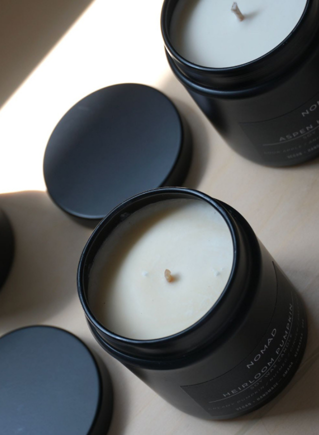 Two soy candles in black jars with labels reading 'Nomad Heirloom Pumpkin', and 'Aspen Hard Cider'