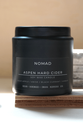 Black candle jar labeled 'Nomad Aspen Hard Cider' on a wooden block with a neutral background