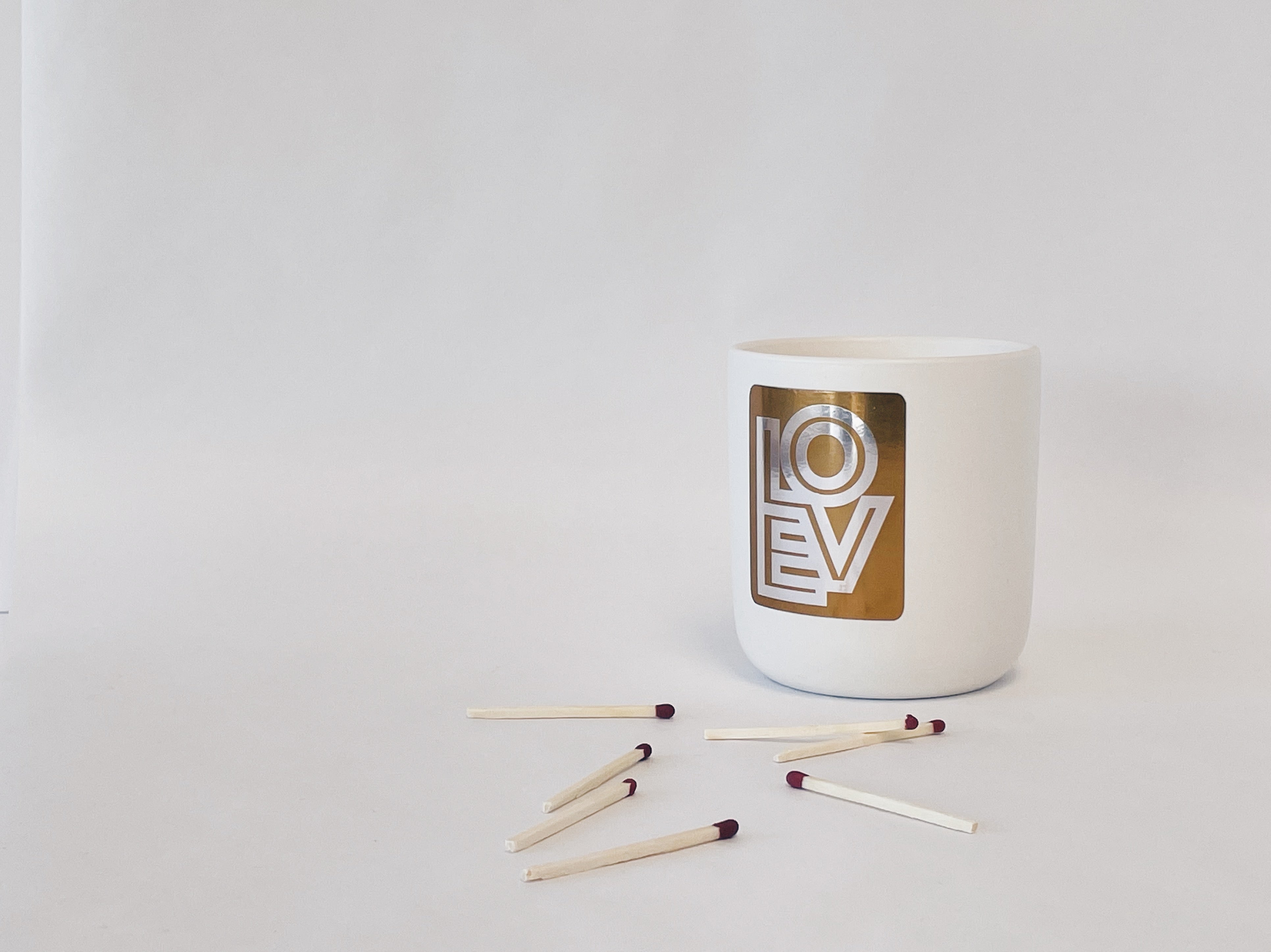 white ceramic candle with silver and gold label that says LOVE, red wood matches scattered in the foreground