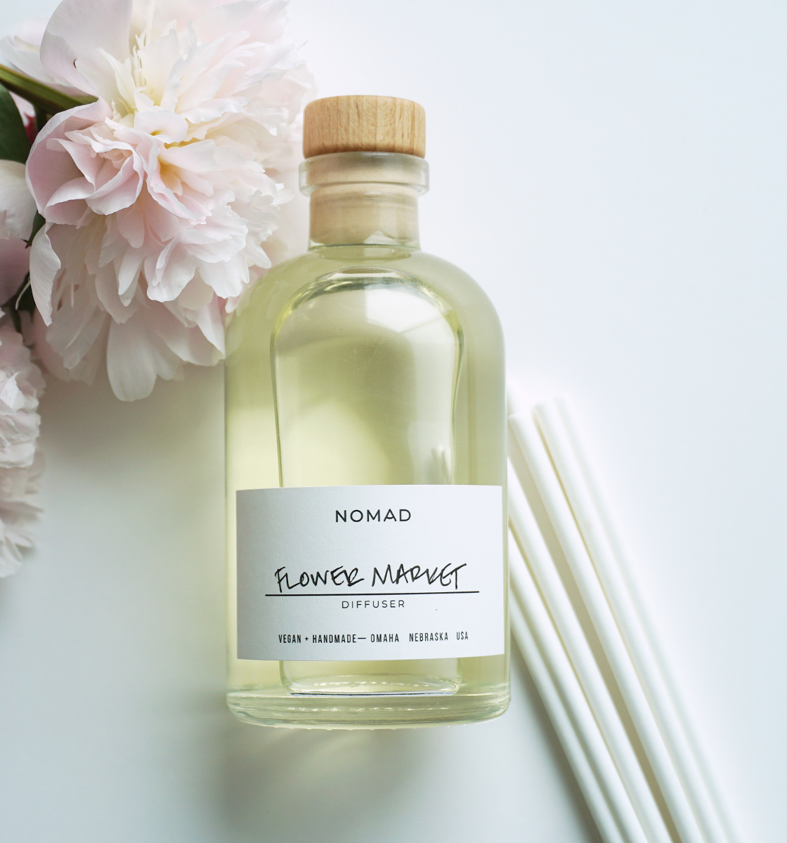 Close up image of Flower Market glass bottle reed diffuser with white label and white reeds with a pink peony on white background